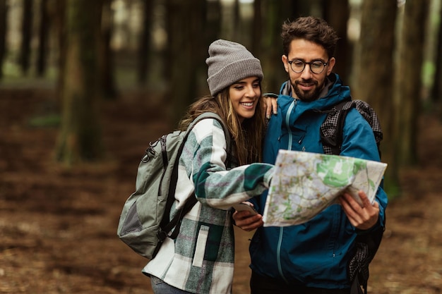 Wesoła młoda para kaukaska w kurtkach cieszy się wakacjami podróżniczymi i patrzy na mapę trasy w lesie