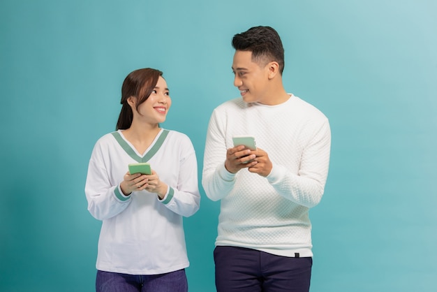 Wesoła młoda para stoi na białym tle nad niebieskim, trzymając smartfony w casual