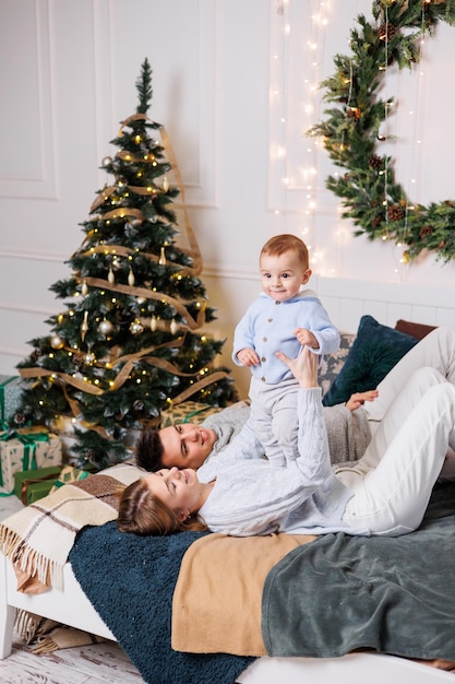 Wesoła młoda para z małym synkiem bawi się na łóżku przy choince Noworoczne wnętrze w sypialni Choinka z zabawkami Świąteczna rodzinna atmosfera