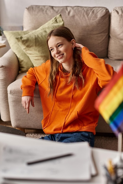 Wesoła nastolatka w pomarańczowym kapturze siedząca w pobliżu kanapy i rozmyta flaga LGBT na pierwszym planie