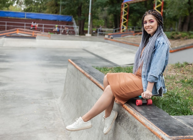 Wesoła niezwykła tysiącletnia kobieta z jasnym makijażem i Afrykanką siedzi na deskorolce w skateparku