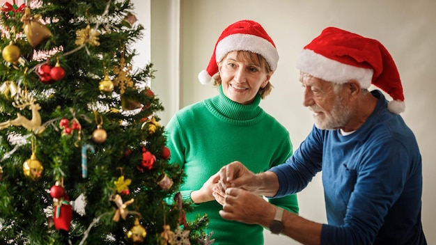 Wesoła para starszych w czapkach Mikołaja uśmiecha się i wiesza bombki na gałęziach drzewa iglastego podczas przygotowań do świętowania Bożego Narodzenia