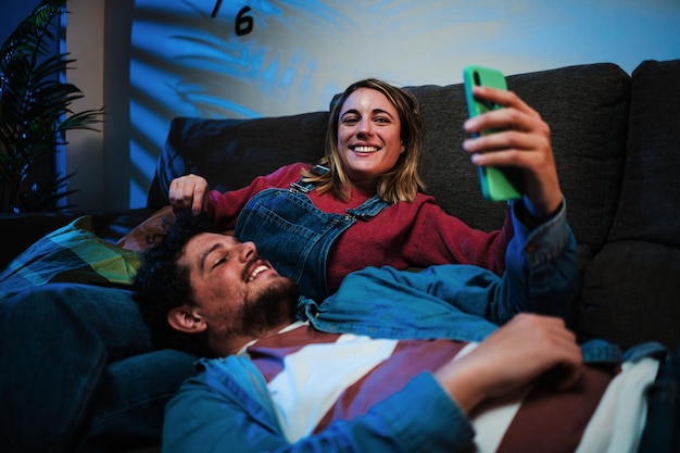 Wesoła para używająca smartfonu odpoczywająca na kanapie w nocy uśmiechnięty chłopak opierający się na swoim