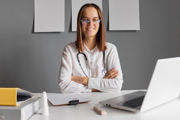 Zdjęcie wesoła, pewna siebie lekarzka pracująca w biurku i uśmiechająca się do kamery w białym płaszczu laboratoryjnym i okularach pozująca ze skrzyżowanymi ramionami robiąc swoją pracę na laptopie w szpitalnym pokoju
