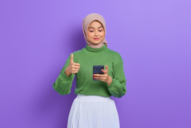 Wesoła piękna Azjatka w zielonym swetrze i hidżabie, korzystająca z telefonu komórkowego i pokazująca kciuk w górze na białym tle na fioletowym tle