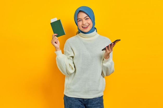 Wesoła piękna Azjatycka kobieta w białym swetrze trzymająca telefon komórkowy i paszport na białym tle na żółtym tle Letnia wycieczka