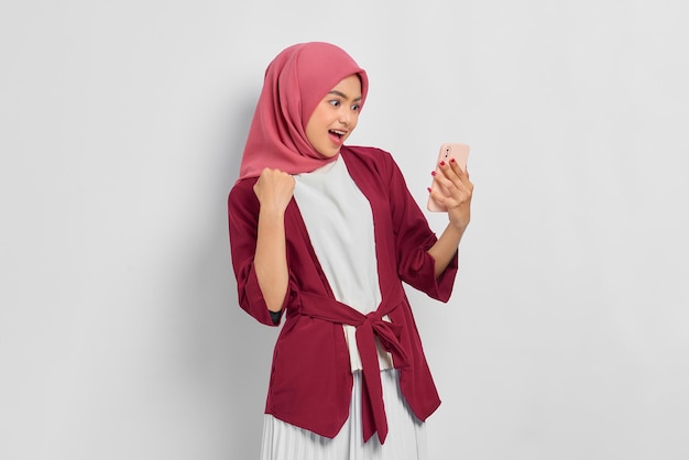 Wesoła piękna Azjatycka kobieta w casualowej koszuli i hidżabie za pomocą telefonu komórkowego, świętując sukces, otrzymując dobre wieści na białym tle nad białym tłem. Koncepcja życia religijnego ludzi