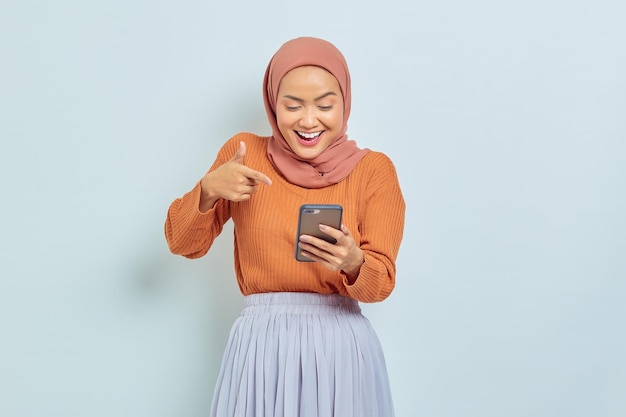 Wesoła piękna Azjatycka Muzułmanka w brązowym swetrze i hidżabie palcem wskazującym na smartfonie na białym tle Koncepcja muzułmańskiego stylu życia