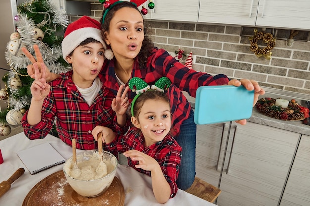 Wesoła piękna latynoska mama z obręczą z poroża ze swoimi uroczymi dziećmi w obręczy elfa i czapce Świętego Mikołaja, wspólnie się bawiąc, gotując świąteczny chleb i robiąc selfie patrząc na kamerę internetową w telefonie