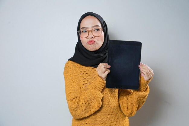 Wesoła, piękna, młoda azjatycka muzułmanka w pomarańczowym swetrze i okularach podczas korzystania z tabletu