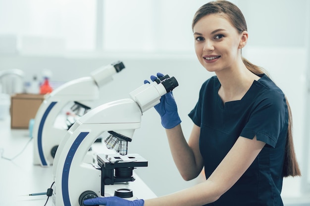 Wesoła pracownica medyczna w mundurze i gumowych rękawiczkach siedzi przy stole z ręką dotykającą mikroskopu