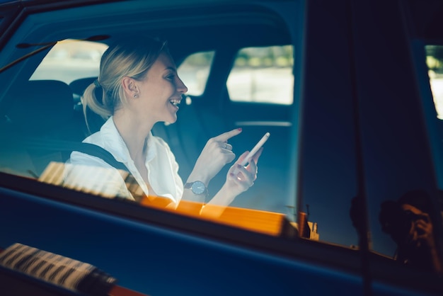 Wesoła profesjonalistka trzymająca w ręku gadżet z telefonem komórkowym, siedząca w samochodzie i śmiejąca się