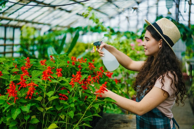 Wesoła rolniczka spryskuje kwitnące kwiaty butelką z rozpylaczem w szklarni