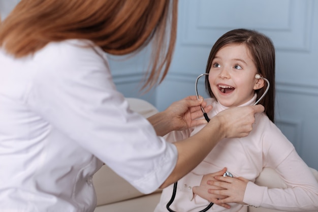 Wesoła śliczna mała dziewczynka trzyma stetoskop i siedzi na kanapie z profesjonalnym lekarzem, wyrażając zadowolenie