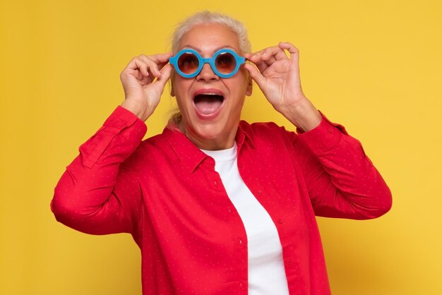 Wesoła stara kobieta nosi modne okulary przeciwsłoneczne odizolowane na żółtym tle śmiejąc się