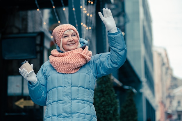Zdjęcie wesoła starsza kobieta spacerująca na świeżym powietrzu w chłodne dni z kartonową filiżanką kawy i machająca ręką, odwracając wzrok z uśmiechem