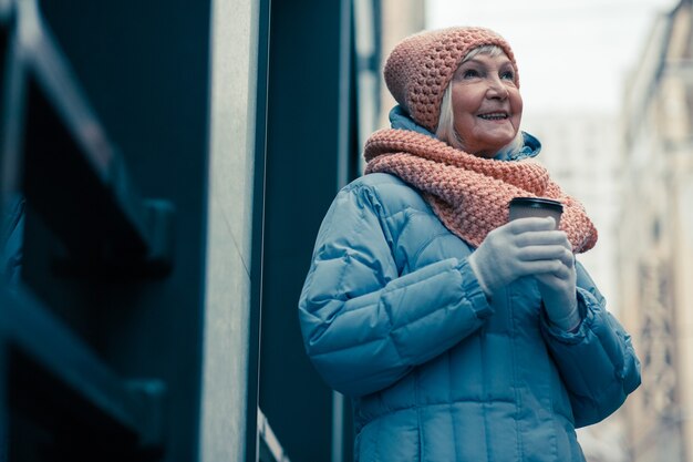 Zdjęcie wesoła starsza kobieta w ciepłym płaszczu zimowym, trzymająca w dłoniach kartonową filiżankę kawy i uśmiechnięta, patrząc w górę