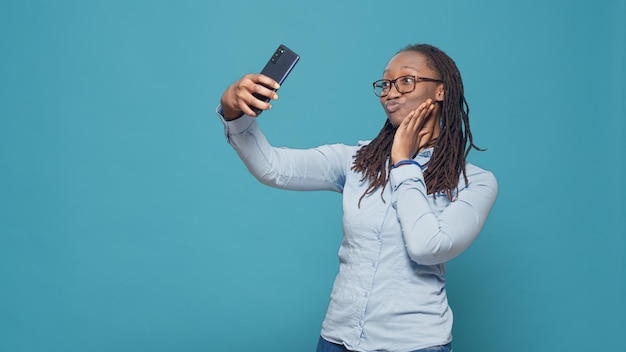 Wesoła szczęśliwa kobieta robienia zdjęć w aplikacji na smartfony, zabawy z aparatem telefonu komórkowego, aby wziąć śmieszne selfie w studio. Naturalny pewnie osoba fotografująca na niebieskim tle.
