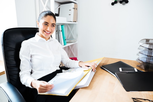 Wesoła uśmiechnięta bizneswoman czyta dokumenty siedząc przy biurku
