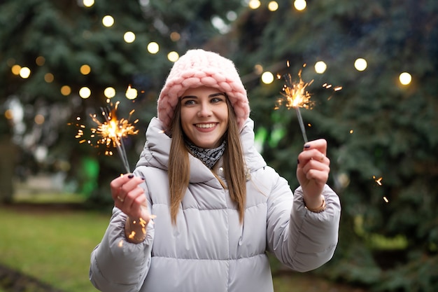 Wesoła uśmiechnięta kobieta bawi się z zimnymi ogniami w pobliżu drzewa noworocznego