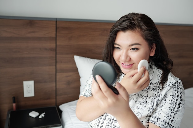 Wesoła Uśmiechnięta Młoda Kobieta Siedzi Na łóżku W Pokoju Hotelowym I Nakłada Kompaktowy Puder, Aby Zmatowić Twarz Przed Spotkaniem Biznesowym Ze Współpracownikami