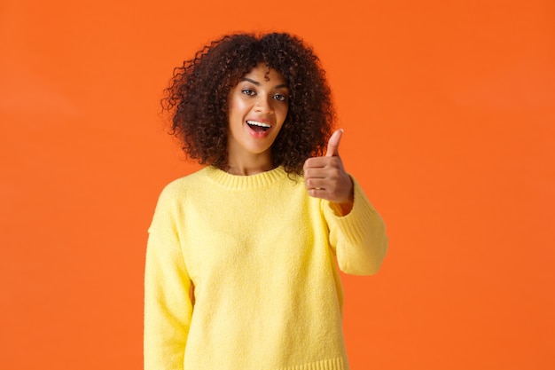 Wesoła wychodząca nowoczesna kobieta Afroamerykanów z kręconymi włosami, pokazująca kciuk w geście aprobaty lub aprobaty, mówiąc wszystko w porządku, tak, uśmiechnięta zadowolona, pomarańczowa ściana
