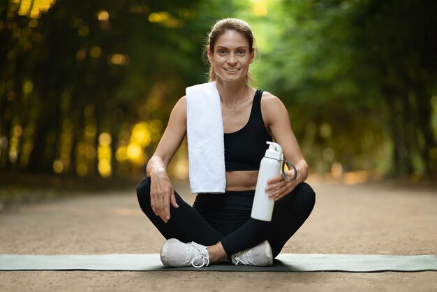 Wesoła wysportowana dama siedzi na macie fitness, trenując na świeżym powietrzu