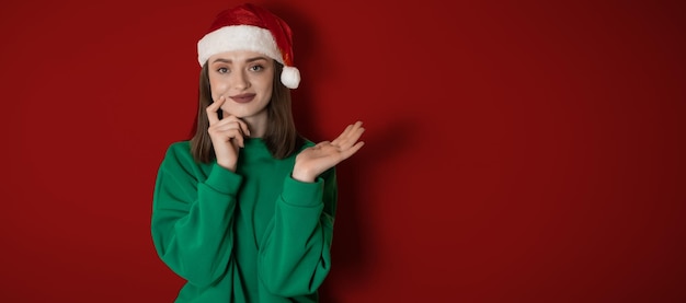 Wesoła zaskoczona młoda kobieta nosi świąteczny sweter Santa hat pozowanie wskazując ręce ramiona na bok wskazuje na obszar roboczy odizolowany na zwykłym pastelowym jasnozielonym tle koncepcja świąteczna szczęśliwego nowego roku 2023