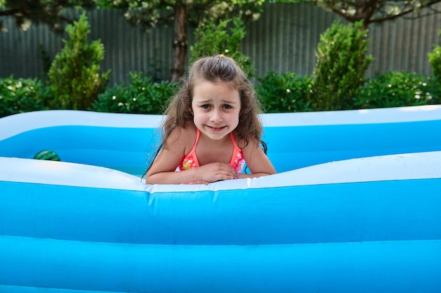 Wesołe dziecko rasy kaukaskiej urocza dziewczynka pływa w nadmuchiwanym basenie i uśmiecha się uroczo patrząc w kamerę