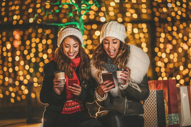Wesołe młode koleżanki surfują w sieci społecznościowej na smartfonie i bawią się w wakacyjną zimową noc na ulicy miasta.
