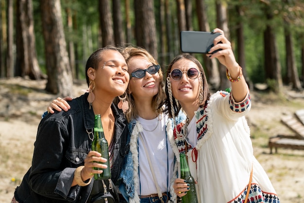 Wesołe młode wieloetniczne kobiety pozujące z piwem do selfie podczas spędzania czasu w leśnym obozie