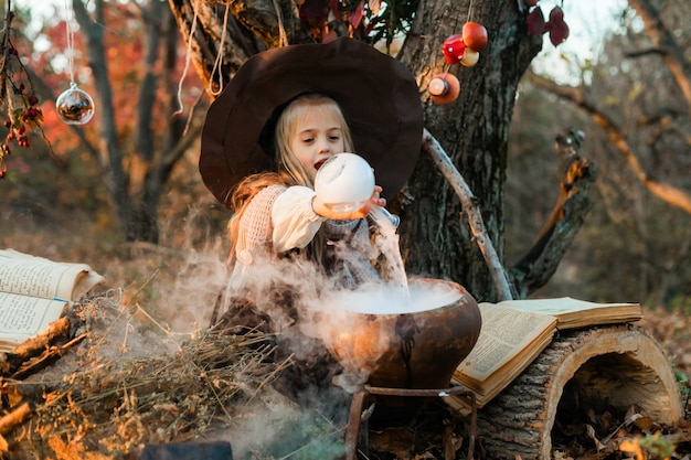 Wesołego Halloween. Słodka dziewczyna w stroju wiedźmy jest w jaskini wiedźmy. Śliczna wesoła mała wiedźma warzy magiczną miksturę. Halloween.