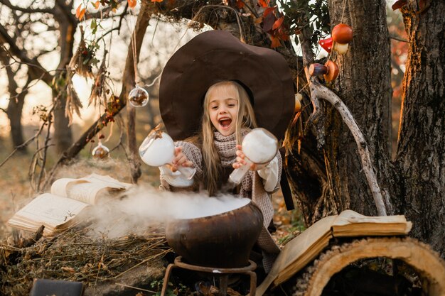 Wesołego Halloween. Słodka dziewczyna w stroju wiedźmy jest w jaskini wiedźmy. Śliczna wesoła mała wiedźma warzy magiczną miksturę. Halloween.