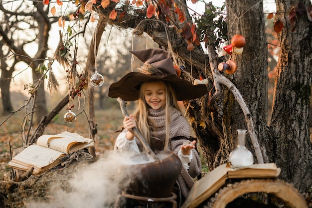 Zdjęcie wesołego halloween. słodka dziewczyna w stroju wiedźmy jest w jaskini wiedźmy. śliczna wesoła mała wiedźma warzy magiczną miksturę. halloween.