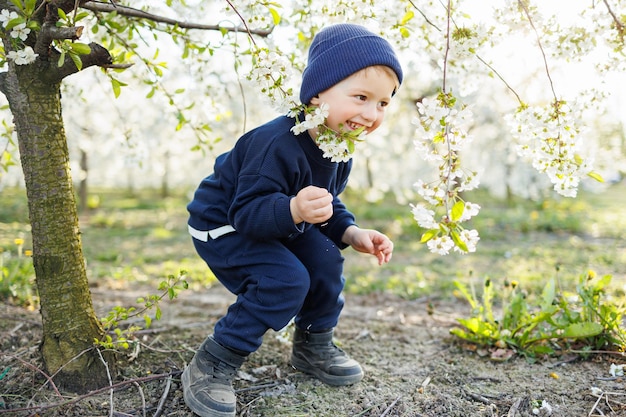 Wesoły 3-latek na spacerze w kwitnącym ogrodzie Mały 3-latek w swetrze i czapce biegnie przez kwitnący ogród Ubranka dla dzieci w wieku 3 lat Emocje dziecięcej radości