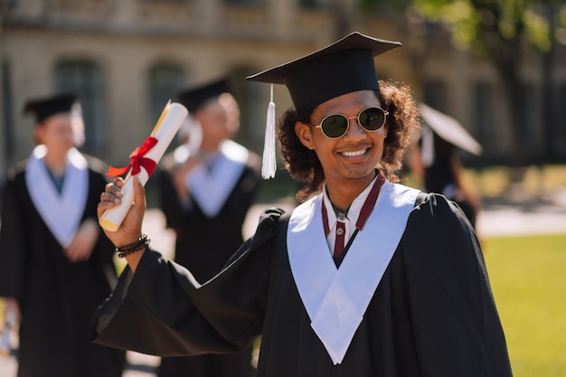 Wesoły absolwent noszący okulary przeciwsłoneczne, podnosząc rękę z dyplomem