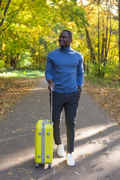 Wesoły African American młody człowiek w stylowe ubrania z walizką spacery po jesiennym parku w ciepły dzień.