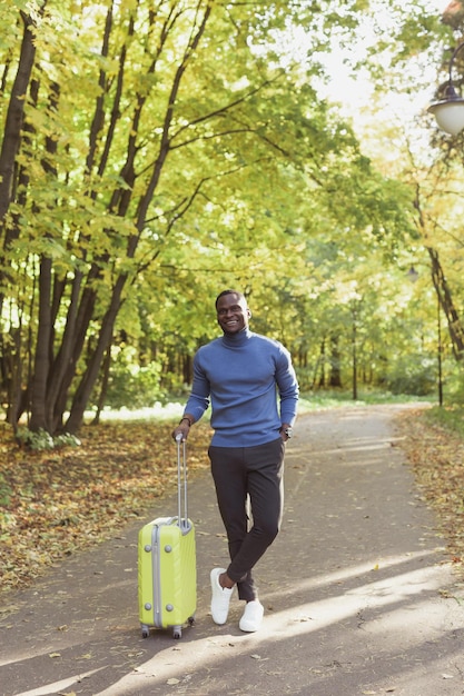 Wesoły afroamerykanin młody człowiek w stylowych ubraniach z walizką spaceruje po wiosennym parku na ciepło