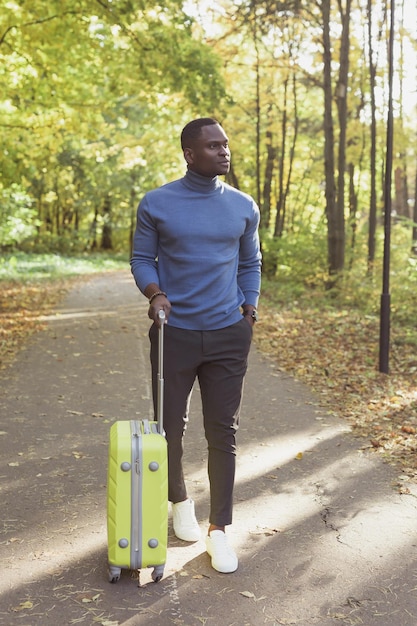 Wesoły afroamerykanin młody człowiek w stylowych ubraniach z walizką spaceruje po wiosennym parku na ciepło