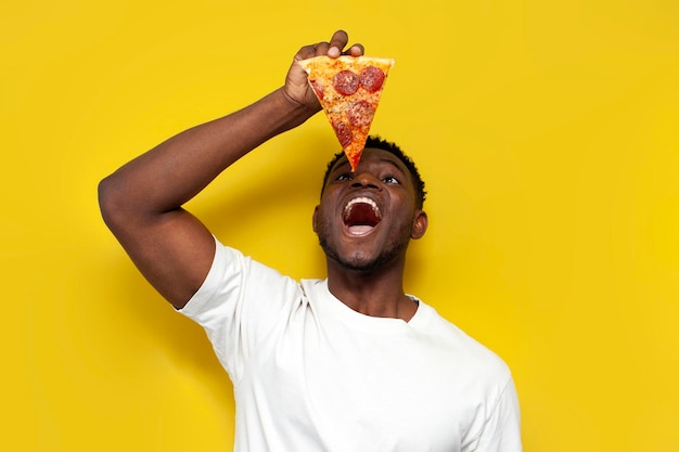 Wesoły Afroamerykanin w białej koszulce trzyma kawałek pizzy z otwartymi ustami.