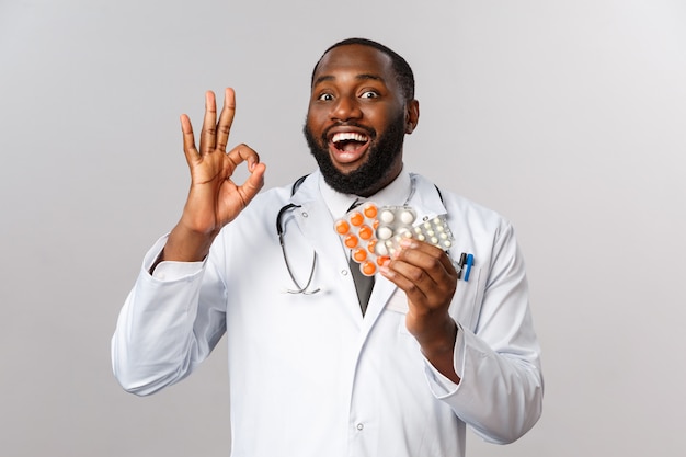 Wesoły afroamerykański przystojny lekarz pokazuje w porządku i medycyny, zapewnia jakość pigułek.