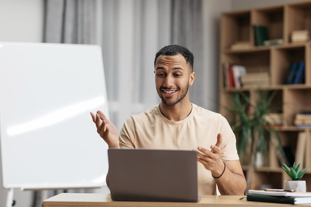 Wesoły arabski nauczyciel patrzący na laptopa i gestykulujący siedzący przy biurku przy tablicy w domu