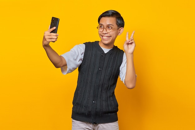 Wesoły azjatycki mężczyzna używający telefonu komórkowego robiący selfie na żółtym tle