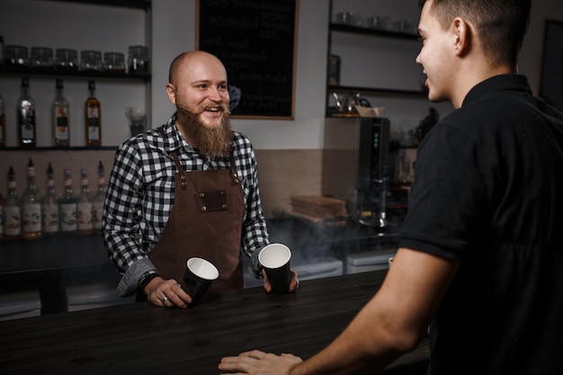 Wesoły barista z brodą sprzedaje kawę kupującemu w barze w nowoczesnej kawiarni