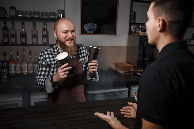 Wesoły barista z brodą sprzedaje kawę kupującemu w barze w nowoczesnej kawiarni