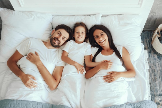 Wesoły brodaty mężczyzna z córką i żoną wyglądający na zrelaksowanych podczas leżenia w łóżku