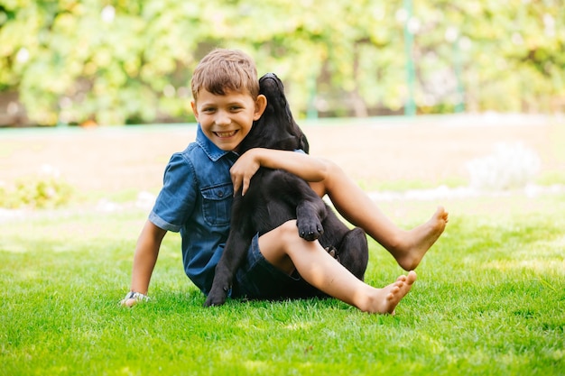 Wesoły chłopak w dżinsowych szortach i koszuli, obejmujący szczęśliwego czarnego szczeniaka Bliskie i ufne relacje między chłopcem a psem, które przetrwają całe życie Spędzanie letnich dni na świeżym powietrzu