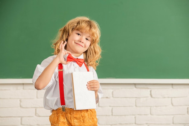 Wesoły chłopiec w muszce w szkolnej klasie z pokazem książki ok gest kopiuj przestrzeń erudycji