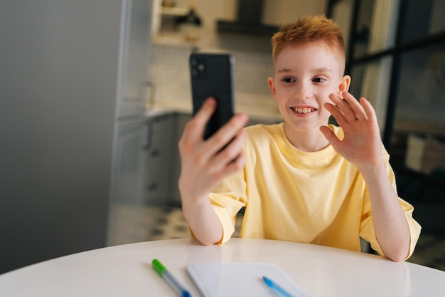 Wesoły chłopiec w szkole trzymający telefon w klasie uczenia się na odległość za pomocą aplikacji mobilnej oglądający lekcję online, rozmowy wideo w aplikacji, robiąc notatki