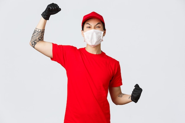 Wesoły entuzjastyczny facet dostawy w czerwonej koszulce i czapce, ubrany w maskę medyczną, rękawice ochronne, podnoszący rękę do śpiewania, powiedz tak lub hura, świętuj sukces, zachęcaj do zamawiania online